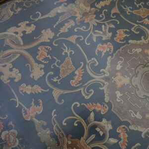 Detailed low-pile rug featuring a dusty blue field with complex cream, taupe, and rust colored mythical patterns.