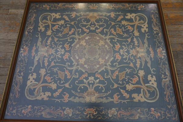 A flat, framed blue rug with an ornate pattern of dragons, phoenixes, and flowers rests on wooden floor planks.