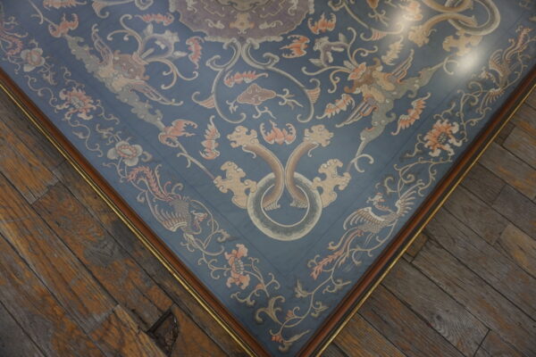 Blue embroidered textile featuring peach and gray mythical motifs, bordered in brown and gold, on worn wood floors.