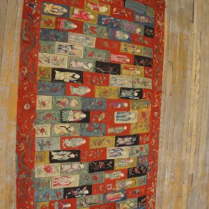 Embroidered multicolored textile rug featuring figures and florals within rectangular blocks, bordered in red, shown on wood flooring.