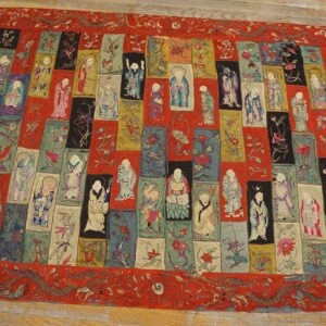 Ornate red textile rug displaying a central design of rectangular panels with multicolored figures and flowers on wood flooring.