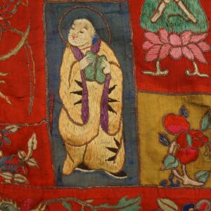 Multicolored stitched textile featuring dense embroidery depicting figures and flowers on red, gold, and blue squares.