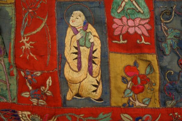 Multicolored stitched textile featuring dense embroidery depicting figures and flowers on red, gold, and blue squares.