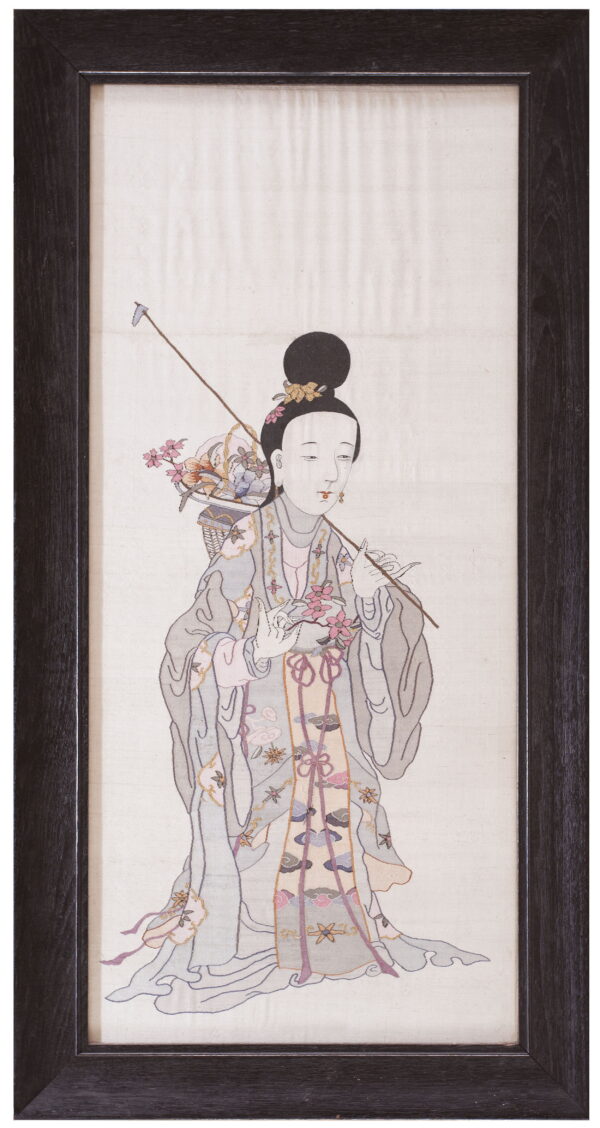 Framed textile art depicting a standing female figure in pale gray and pink robes holding flowers.