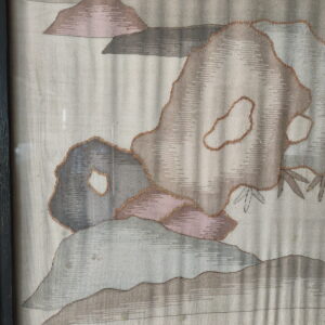 Pale woven textile framed behind glass, featuring abstract shapes in muted gray, pink, and copper outlines.