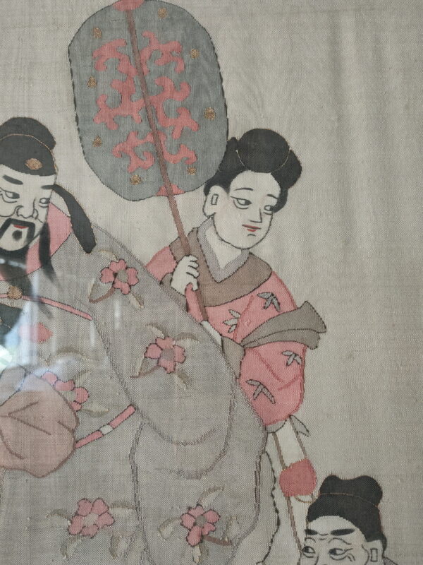 Textile depicting stylized figures, flowers, and a fan, rendered in dusty pink, gray, black, and gold threads.