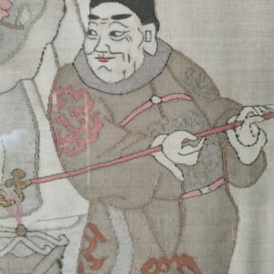 Flat woven textile depicting a figure in taupe and cream robes with pink and gold accents.