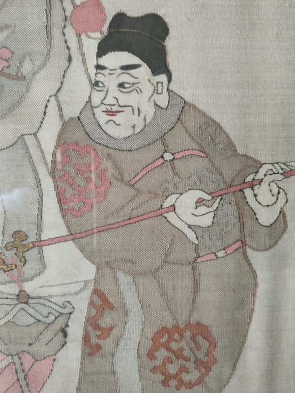 Flat woven textile depicting a figure in taupe and cream robes with pink and gold accents.