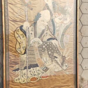 Large embroidered tapestry artwork depicting two figures on a gold background, framed and resting on gray pavement.