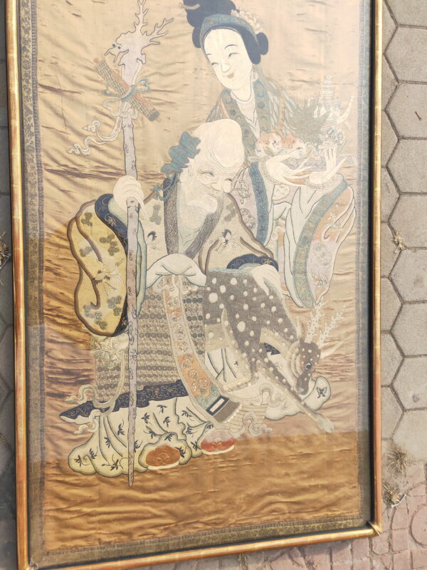 Large embroidered tapestry artwork depicting two figures on a gold background, framed and resting on gray pavement.