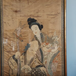 Detailed embroidered textile featuring two figures in tan, blue, and white, framed against a light blue wall.