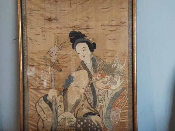 Detailed embroidered textile featuring two figures in tan, blue, and white, framed against a light blue wall.