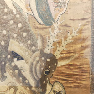 A detailed embroidered textile shows a spotted deer head and antlers on a shimmering tan and brown background.