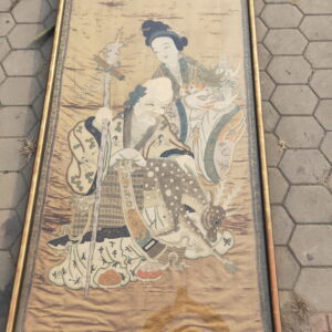 Framed vertical textile with traditional figures and a deer, lying on gray hexagonal outdoor pavers.