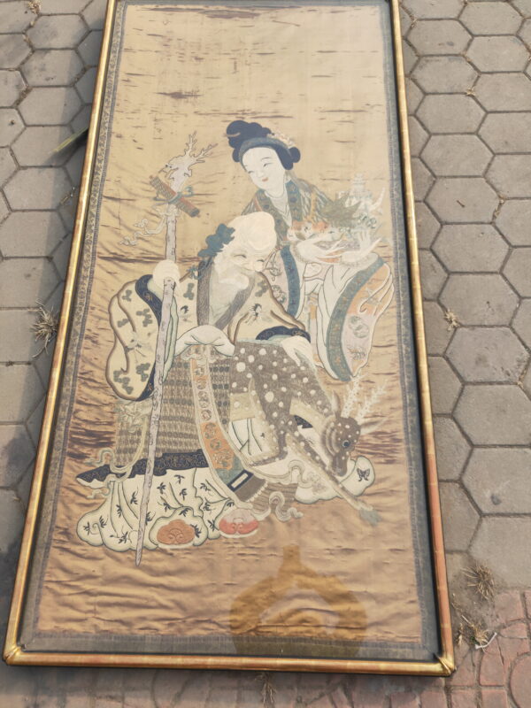 Framed vertical textile with traditional figures and a deer, lying on gray hexagonal outdoor pavers.