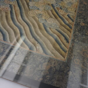 Framed textile corner with wavy blue and tan stripes, cream cloud motifs, and a dark floral border under glass.