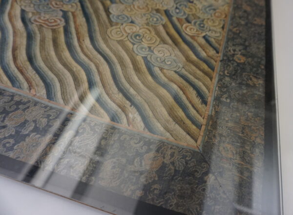 Framed textile corner with wavy blue and tan stripes, cream cloud motifs, and a dark floral border under glass.