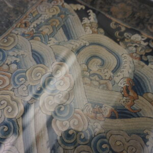 Close-up of a framed textile with fine stitching showing swirling blue waves and stylized clouds.