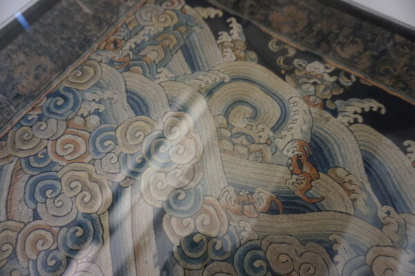 Close-up of a framed textile with fine stitching showing swirling blue waves and stylized clouds.