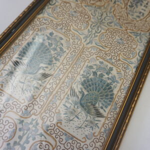 Detailed view of framed blue and white textile featuring peacock figures and gold embroidered borders.