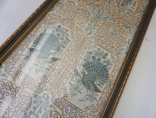 Detailed view of framed blue and white textile featuring peacock figures and gold embroidered borders.