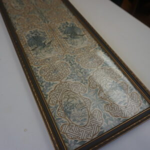 Ornate framed textile runner with blue and cream peacock embroidery and gold geometric patterns rests on a white surface.