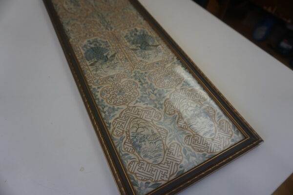 Ornate framed textile runner with blue and cream peacock embroidery and gold geometric patterns rests on a white surface.