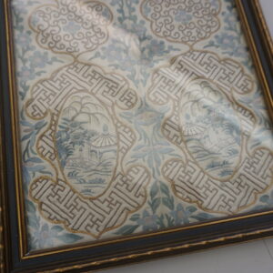 Framed flat textile with intricate embroidery showing pale blue flowers, geometric borders, and small landscape scenes.