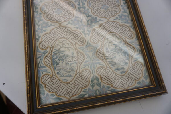Framed flat textile with intricate embroidery showing pale blue flowers, geometric borders, and small landscape scenes.