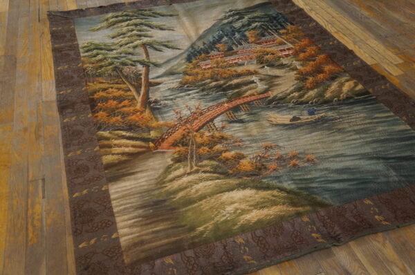 Detailed landscape rug with a river, green trees, orange foliage, and a dark patterned border on wood flooring.