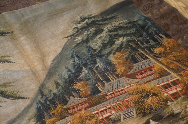 Textured textile featuring a mountainous scene with dense blue trees, orange foliage, and tiered red buildings.