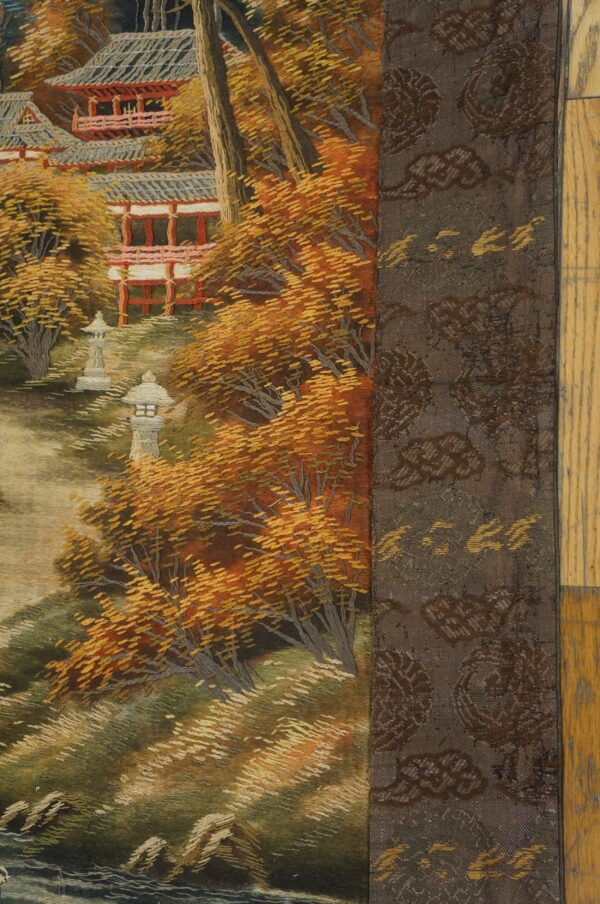 Embroidered textile featuring a detailed autumn scene with red pagodas and vibrant orange trees, adjacent to a dark patterned border.