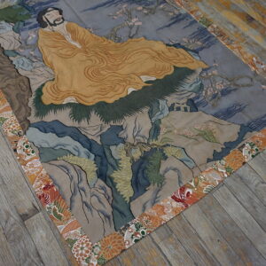 A pictorial rug depicting a figure in yellow on a landscape of gray-blue rocks, framed by an orange floral border.