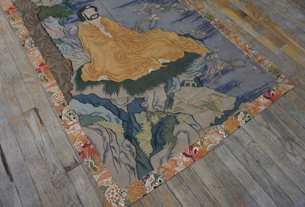 A pictorial rug depicting a figure in yellow on a landscape of gray-blue rocks, framed by an orange floral border.