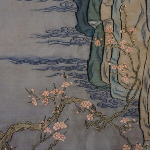 A draped low pile rug displays pink blossoms, teal mountains, and blue waves beside patterned gold textiles.