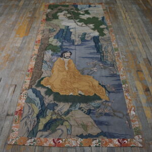 Narrow tapestry featuring a seated figure in yellow robes and a multicolored floral border on distressed wood flooring.