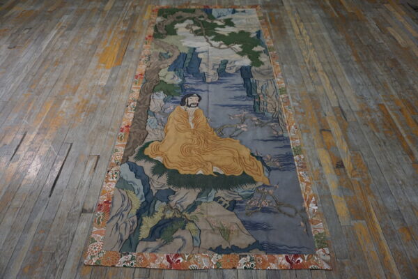 Narrow tapestry featuring a seated figure in yellow robes and a multicolored floral border on distressed wood flooring.