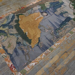 Artistic rug showing a gold-robed figure and blue landscape, finished with an orange floral border.