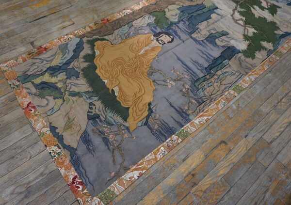 Artistic rug showing a gold-robed figure and blue landscape, finished with an orange floral border.