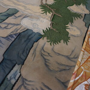 Detailed rug shows blue mountains and a green pine branch beside ornate orange fabric on wood.