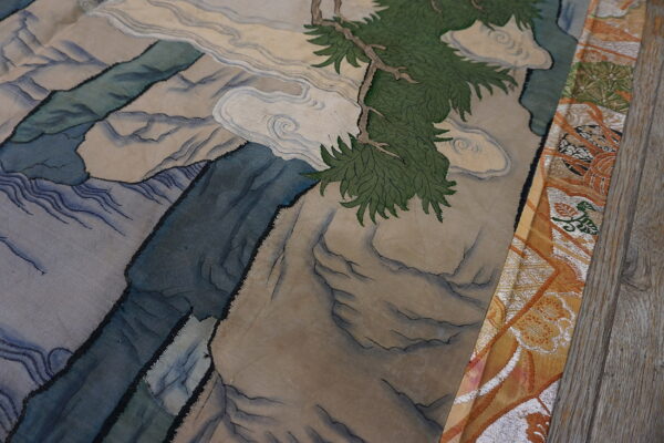 Detailed rug shows blue mountains and a green pine branch beside ornate orange fabric on wood.