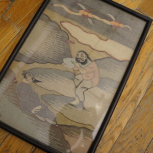 Framed, muted gray and beige pictorial textile art featuring two figures lies on distressed hardwood planks.