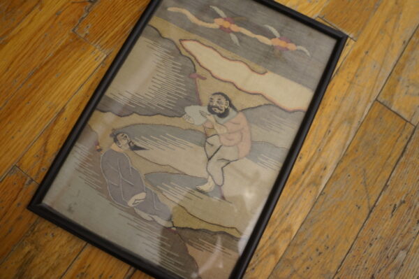 Framed, muted gray and beige pictorial textile art featuring two figures lies on distressed hardwood planks.