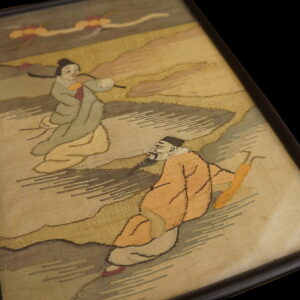 A framed textile art piece displays two figurative characters in orange and sage robes on a muted beige ground.