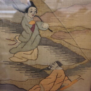 Flat woven textile framed in black, depicting two figures in gray, beige, and gold tones.