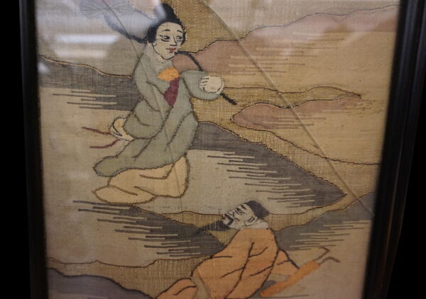 Flat woven textile framed in black, depicting two figures in gray, beige, and gold tones.