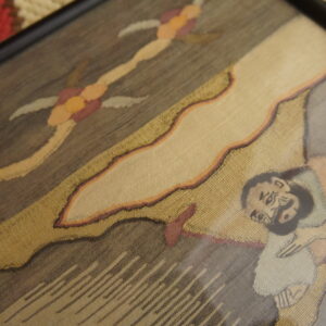 Framed, fine-weave textile featuring figures in gold and brown resting on a thick, red woven rug.