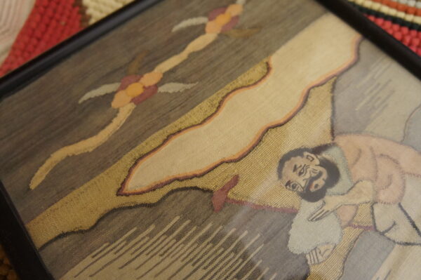 Framed, fine-weave textile featuring figures in gold and brown resting on a thick, red woven rug.
