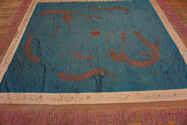 Square teal textile featuring copper-colored embroidered dragons, wide beige floral borders, and heavy magenta fringe.