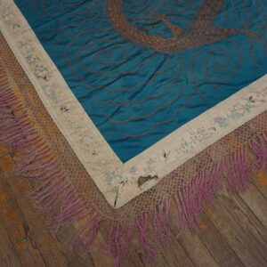 Embroidered blue rug corner featuring brown stitching, a floral border, and long mauve fringe over wood flooring.